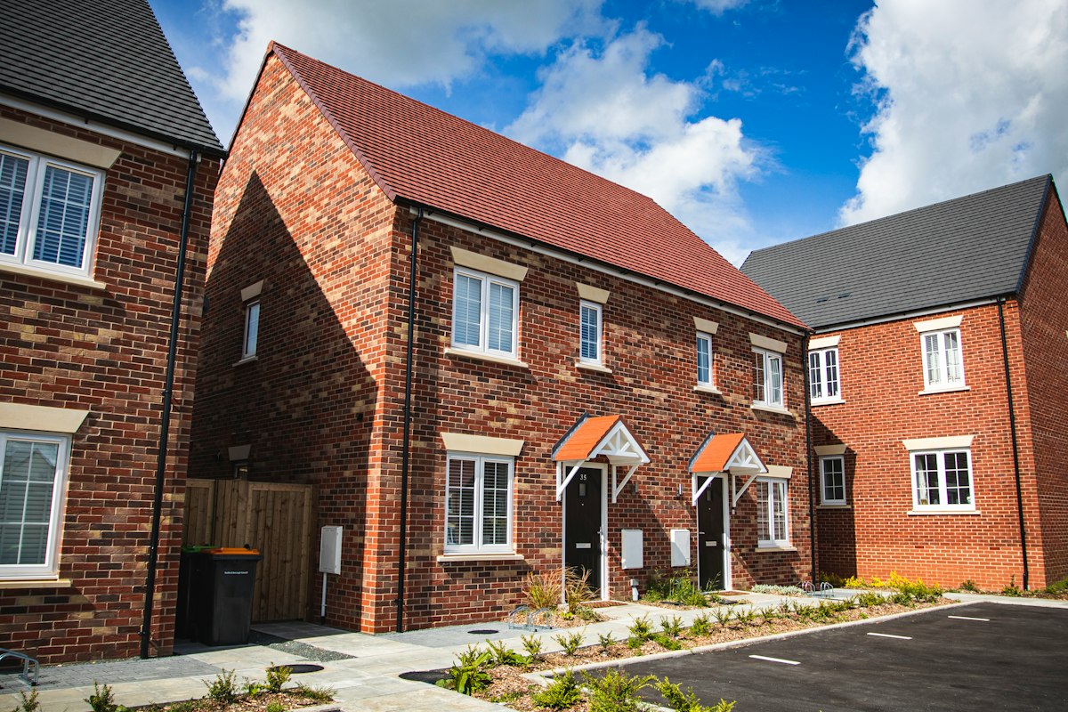 A pair of newly built red-brick semi-detached houses with small canopies over the front doors, beside other new brick homes and a freshly surfaced parking area.