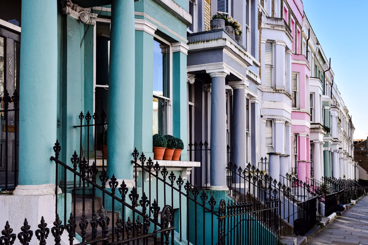 A row of pastel-painted Victorian terraced houses with columned porches and black iron railings along a London street.