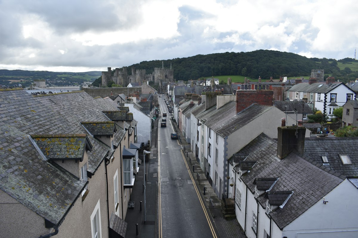 A street of slate-roofed terraced houses in Conwy, North Wales, with Conwy Castle and wooded hills in the background.
