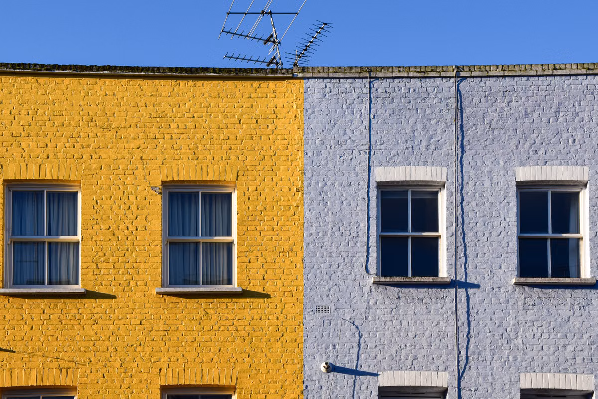 The upper floors of two terraced houses with painted brick, one yellow and one pale blue, with sash windows and a TV aerial against a blue sky.