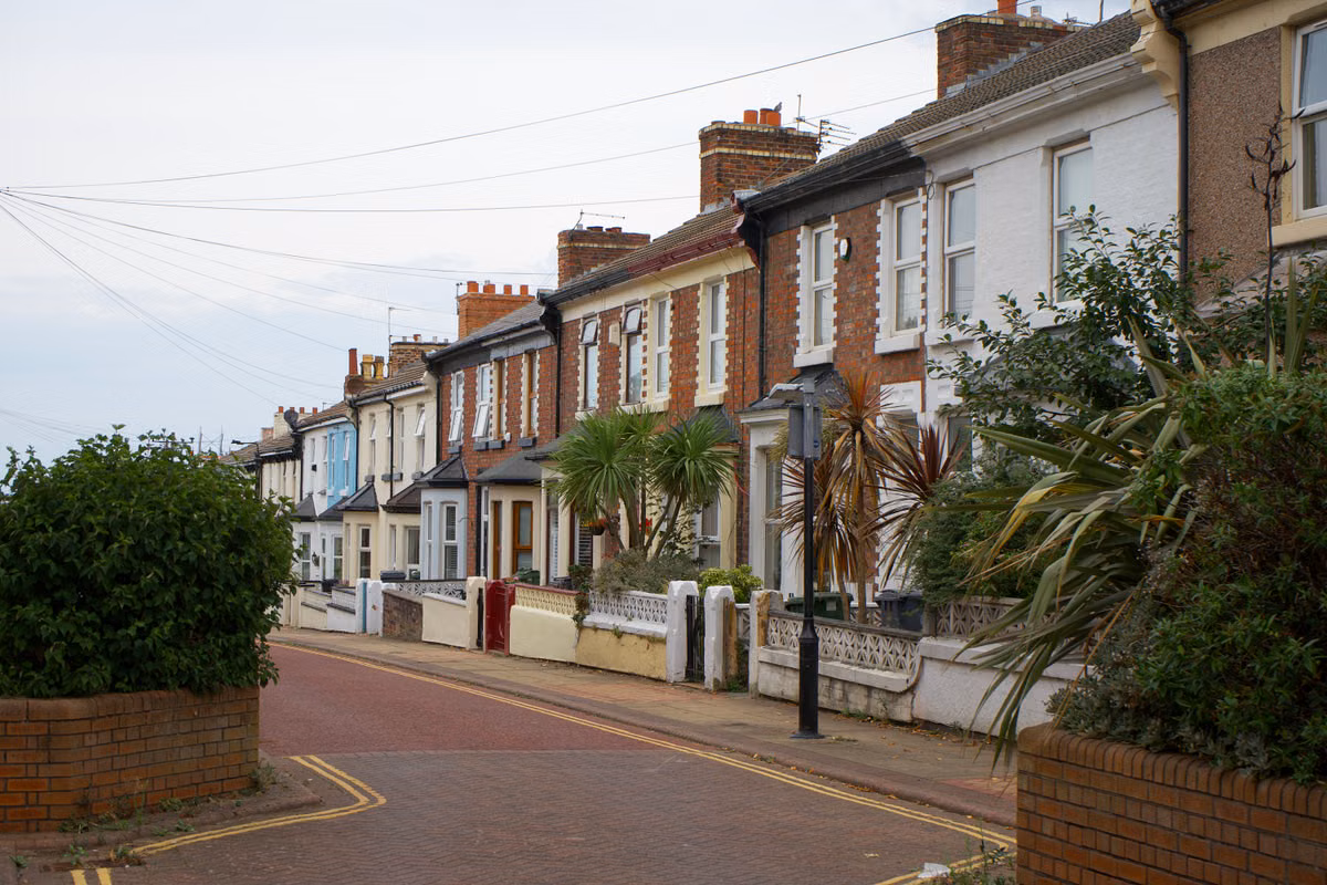 A residential street of two-storey red brick and painted terraced houses with small front gardens, low walls and planting.
