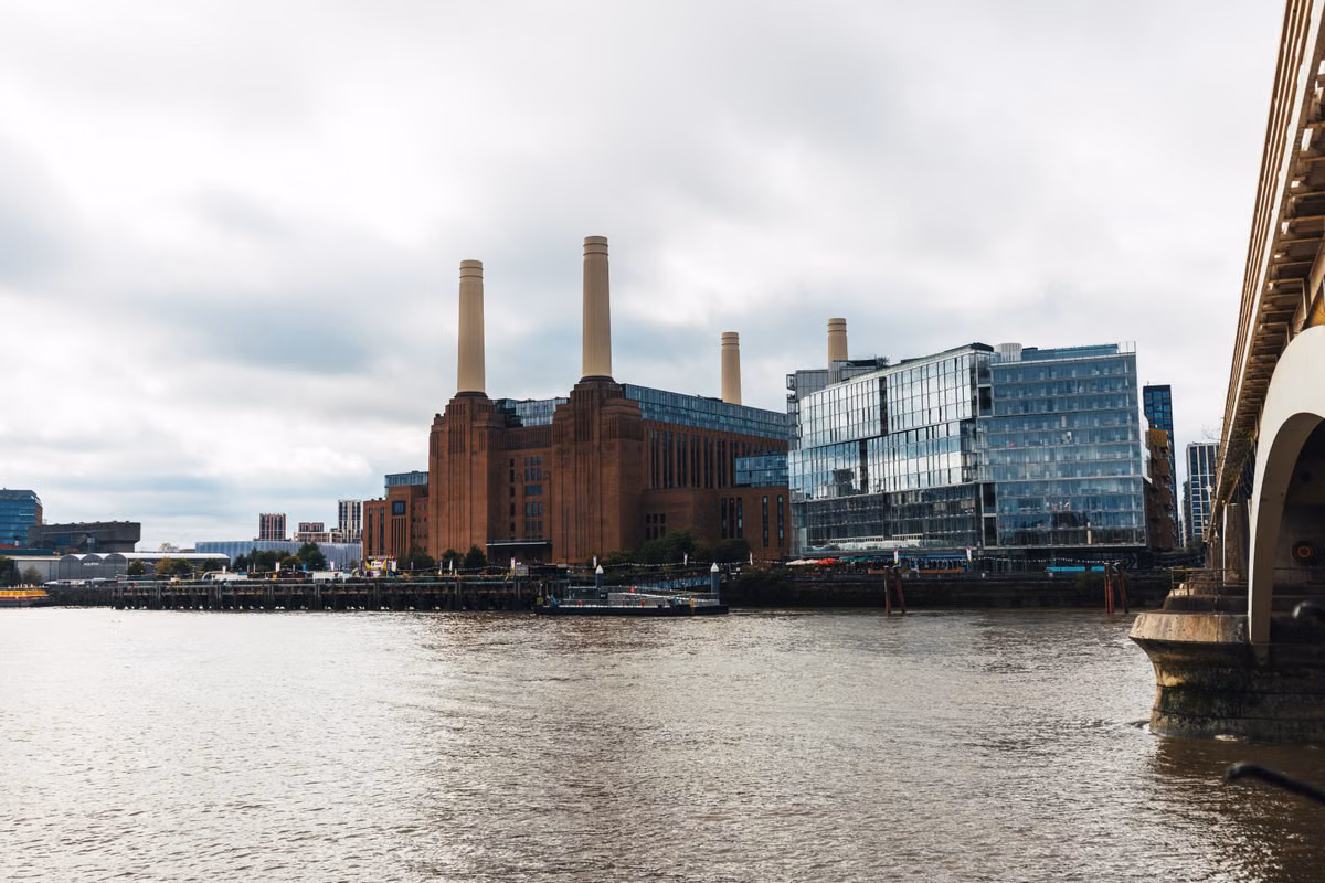 Battersea Power Station's brick building and four chimneys beside newer glass office buildings, seen across the River Thames on an overcast day with a bridge at the right.